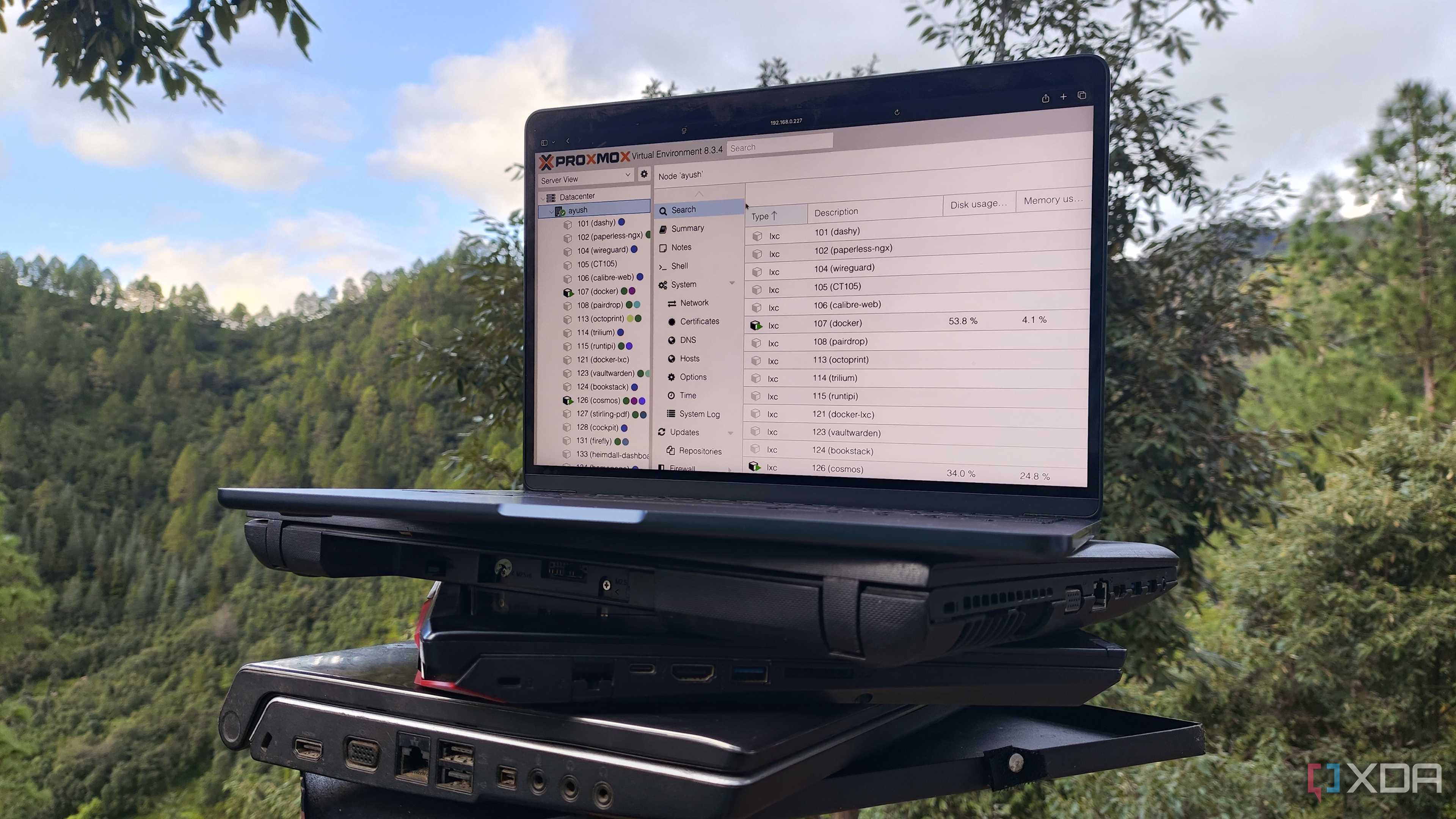 Multiple old laptops, with a MacBook displaying the Proxmox UI resting on top of them