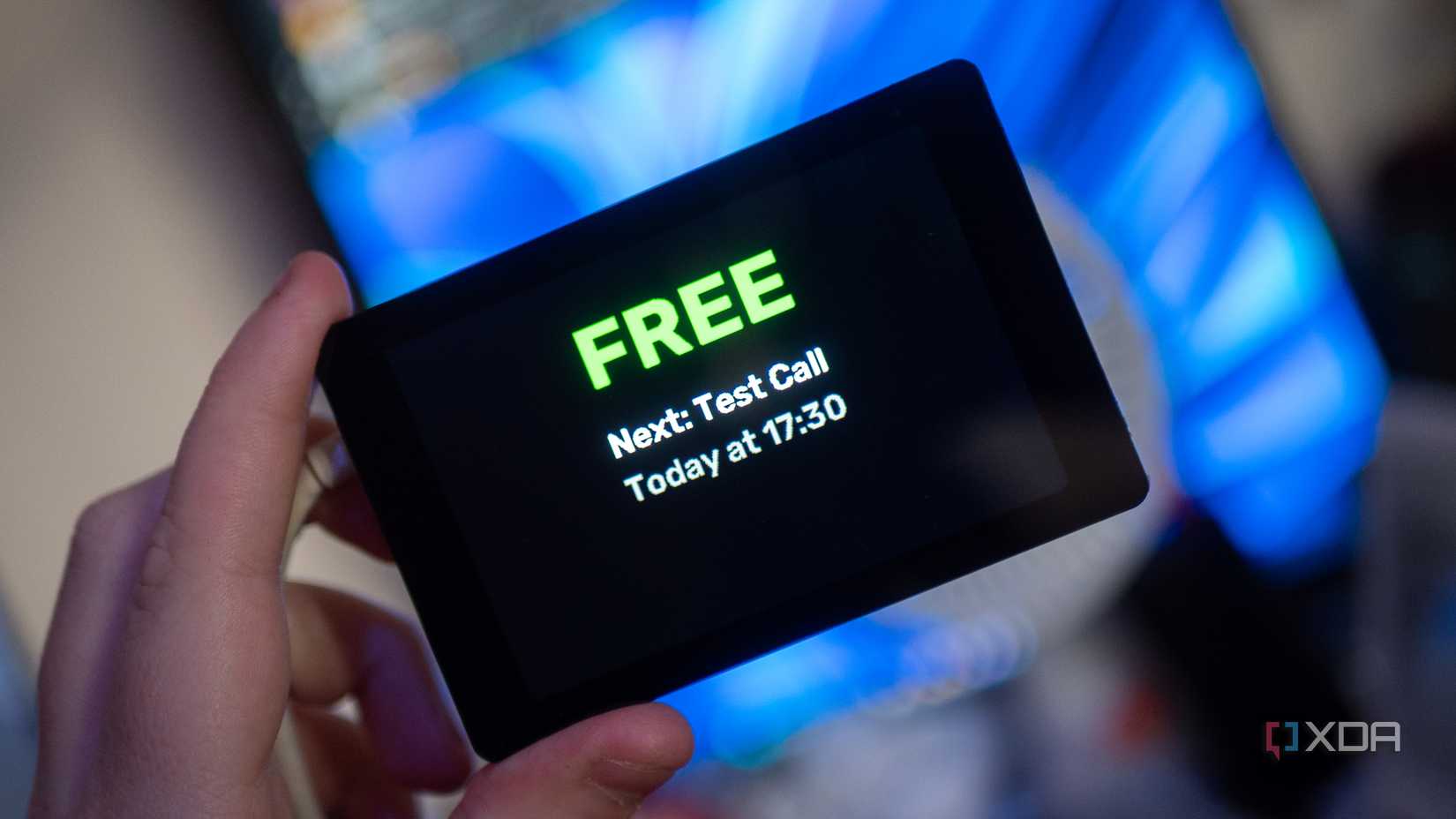 Hand holding an ESP32 display, showing “FREE” and “Next: Test Call Today at 17:30.” Blue light background.