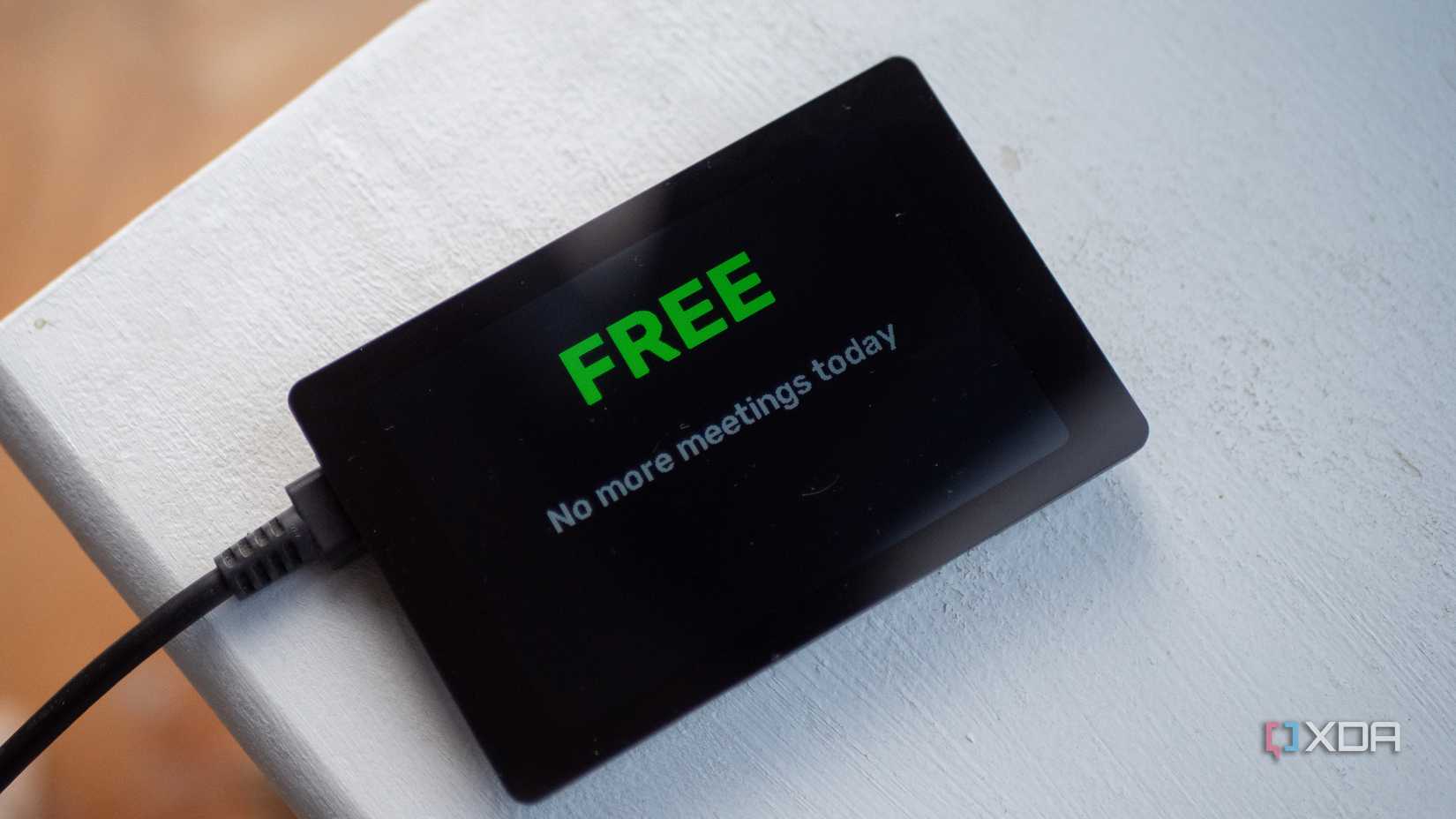 Small ESP32 display on a white surface reads "FREE No more meetings today." Cable connected to left side.
