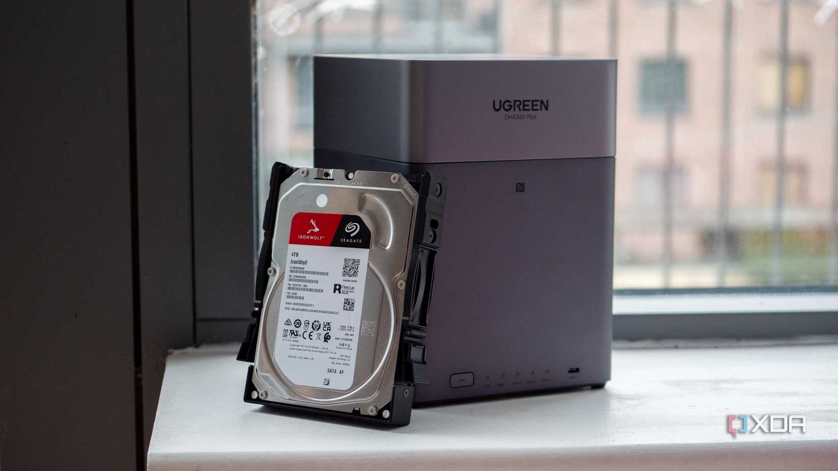 UGREEN DH4300 Plus and a Seagate Iron Wolf hard drive sit on a ledge in front of a window overlooking buildings. 