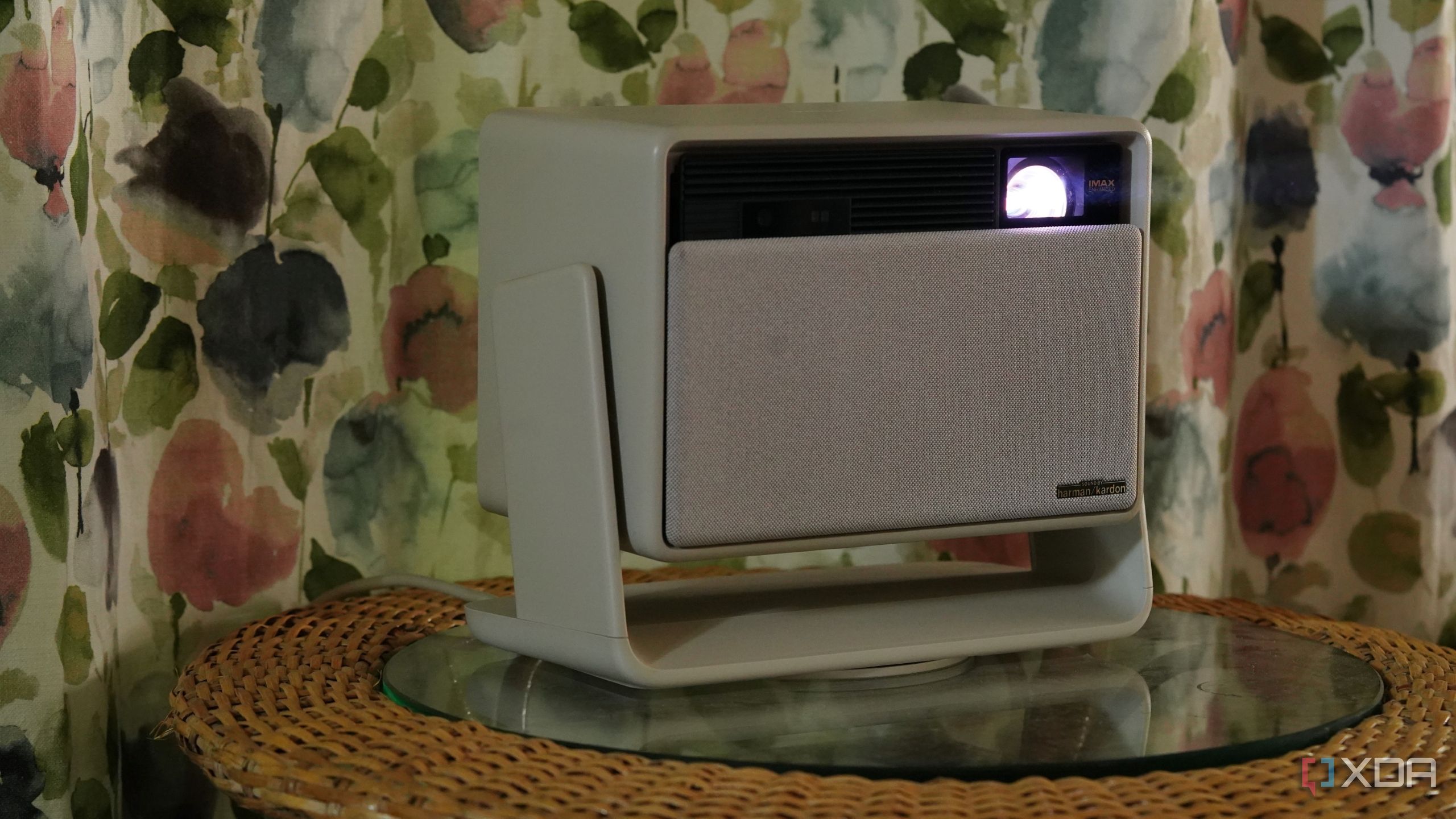 A beige home theater projector on a cane table with floral curtains behind it