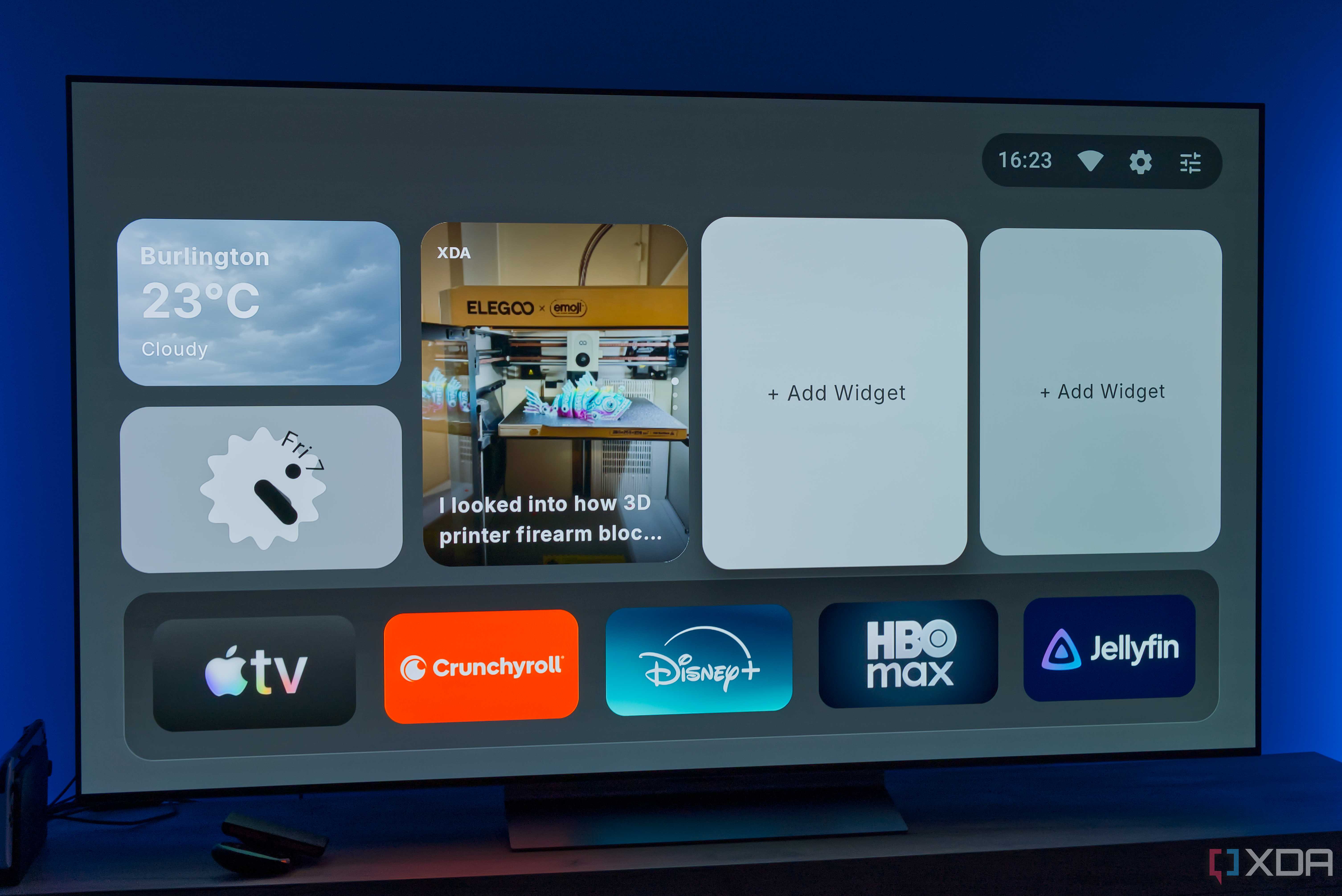 A TV displaying the homepage for AT4K Launcher for Android TV, with a small weather widget, a small analog clock widget, and one full-size RSS feed widget for XDA. Two widget slots are left open