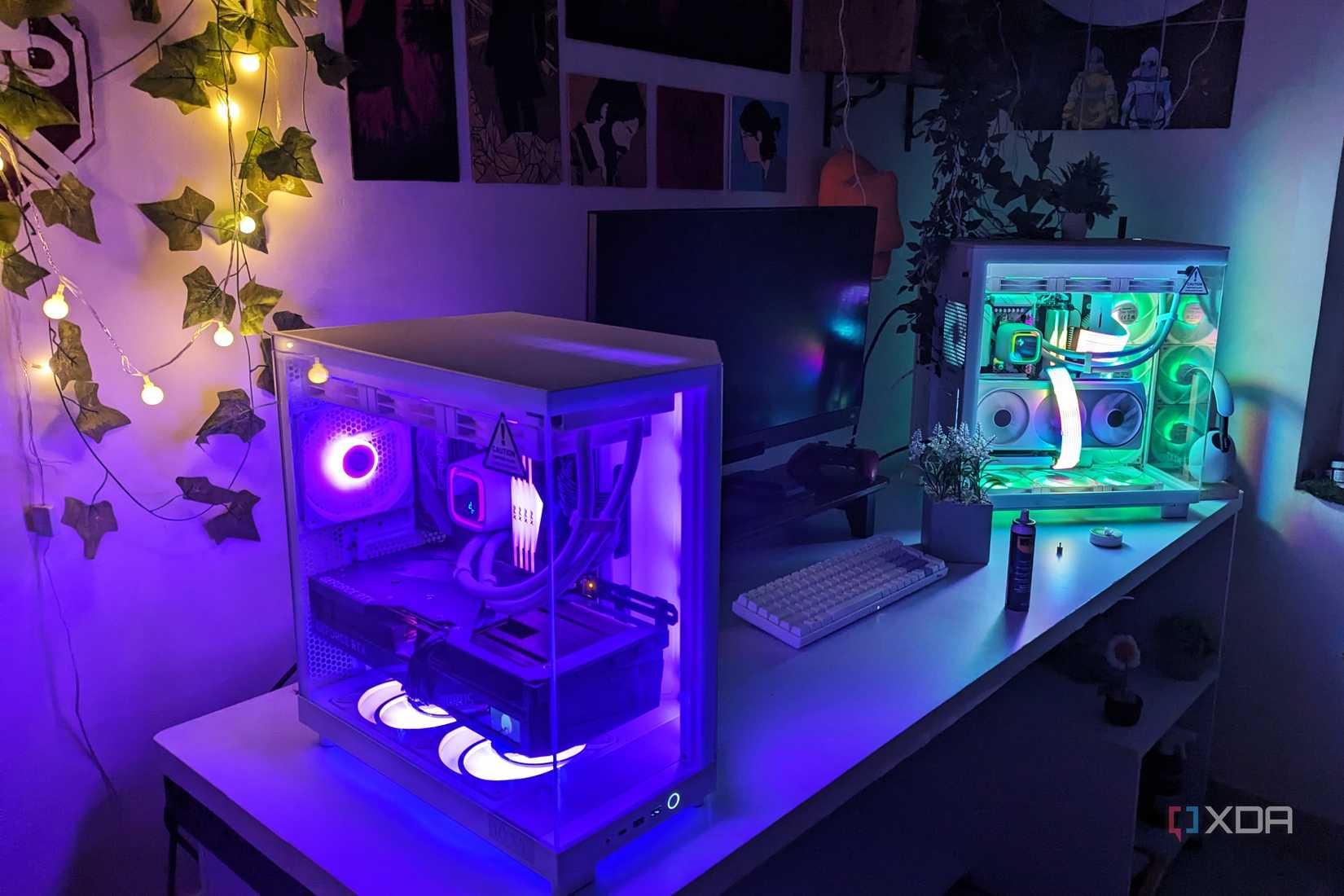Two gaming PCs with RGB lighting next to a monitor on a white desk in dim lighting
