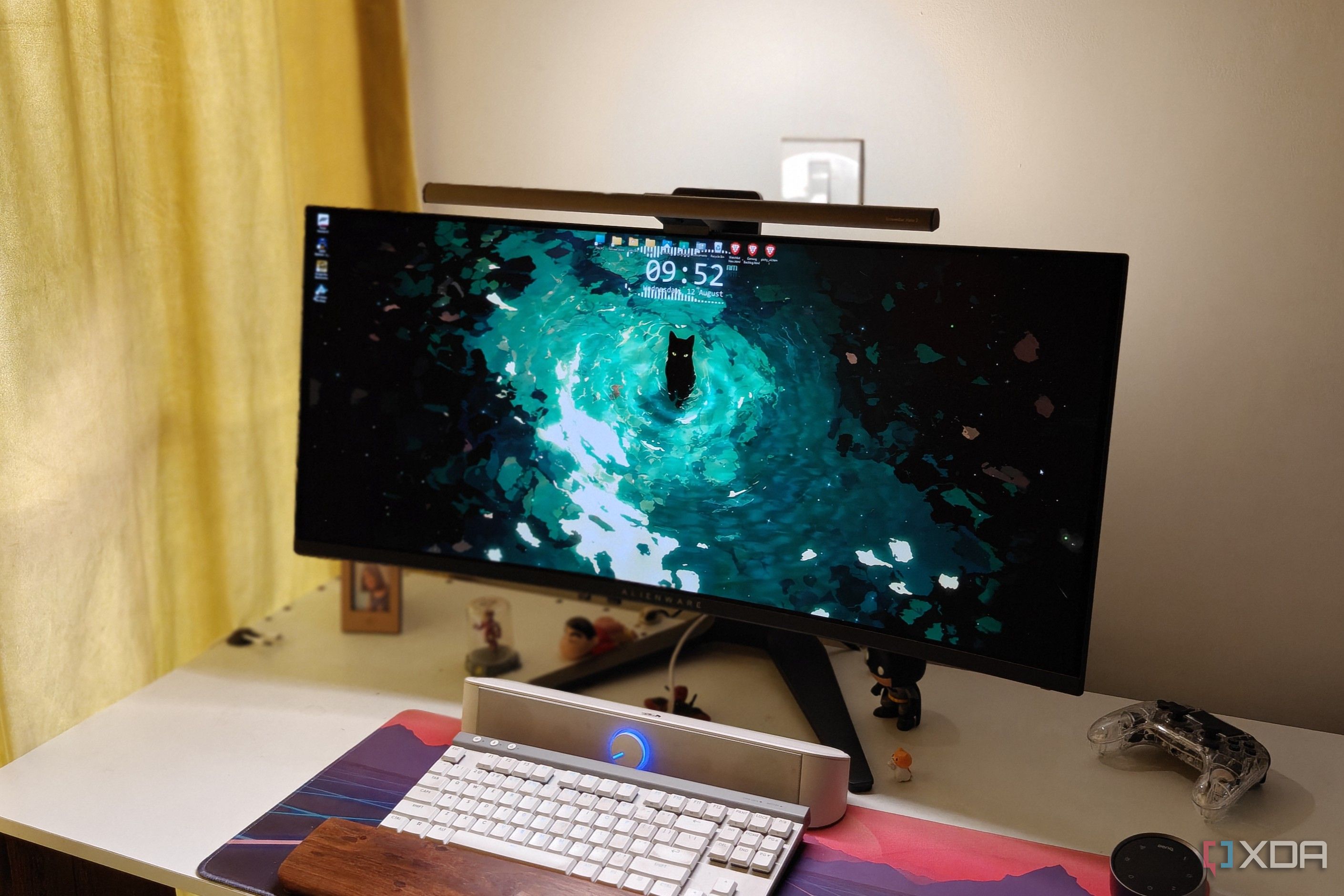 An ultrawide monitor with a light bar on a white desk with a keyboard and soundbar