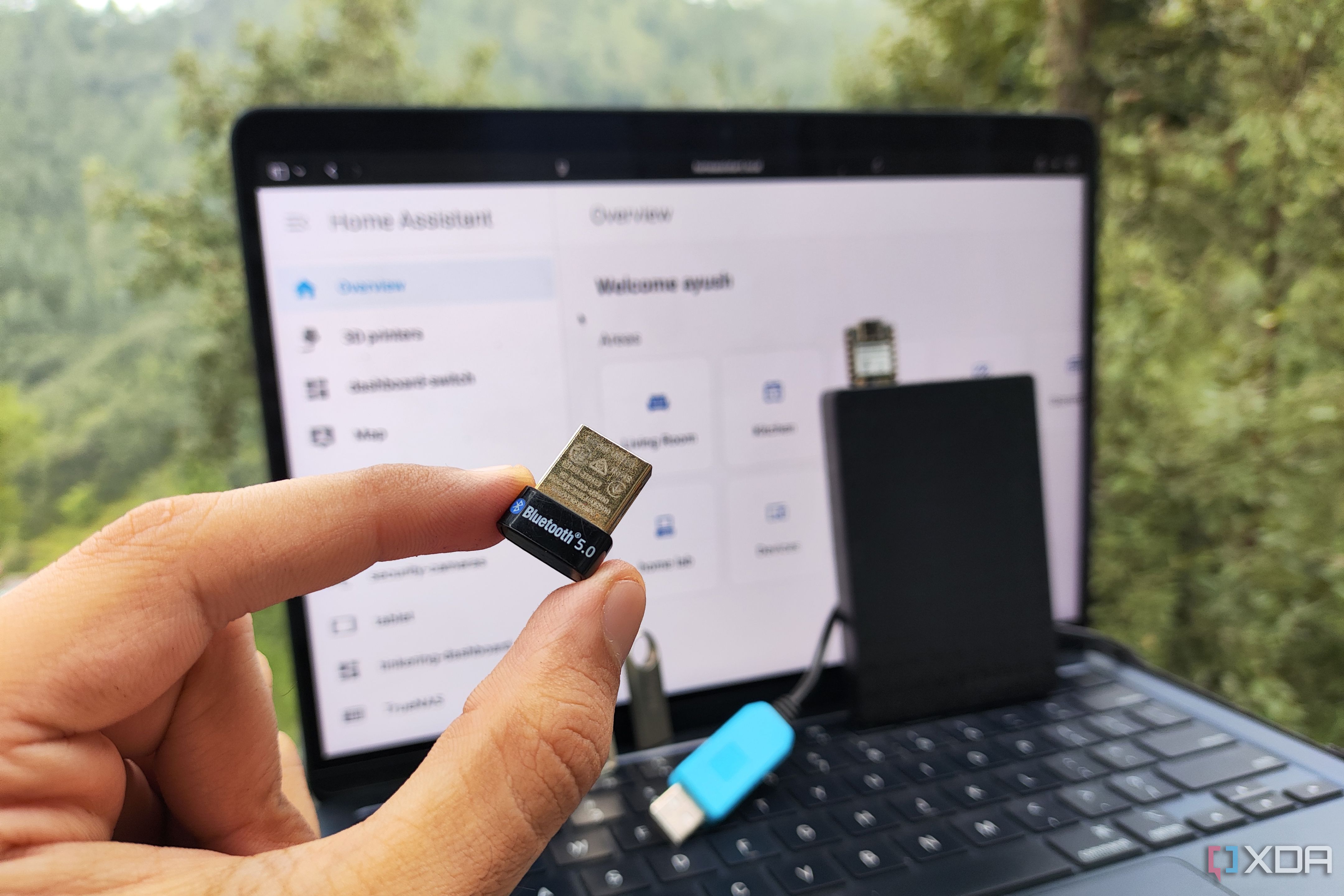 A person holding a Bluetooth dongle and USB devices in front of a macOS running the Home Assistant web UI