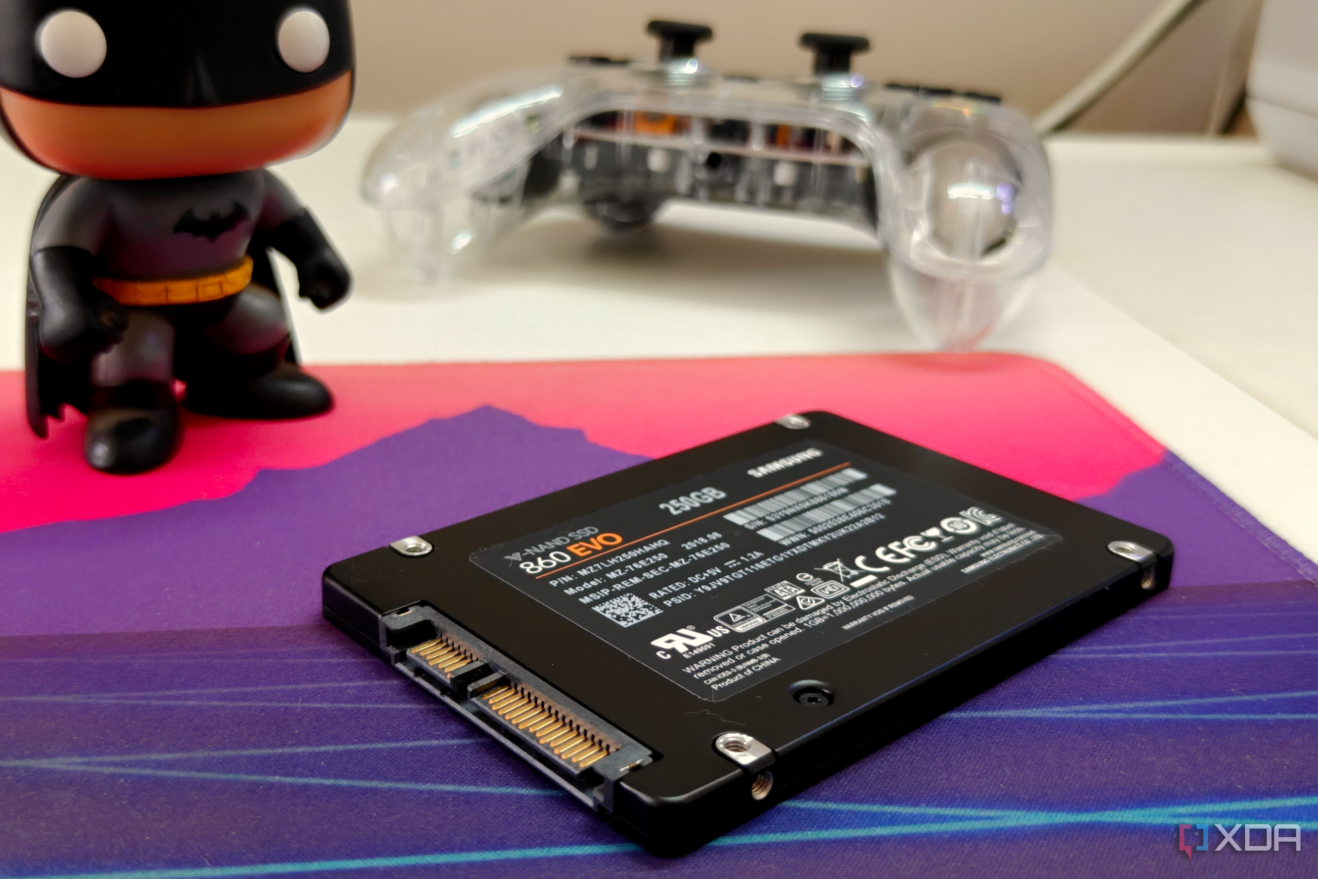 Close-up of a black SATA SSD on a deskmat next to a Batman figurine and a gaming controller