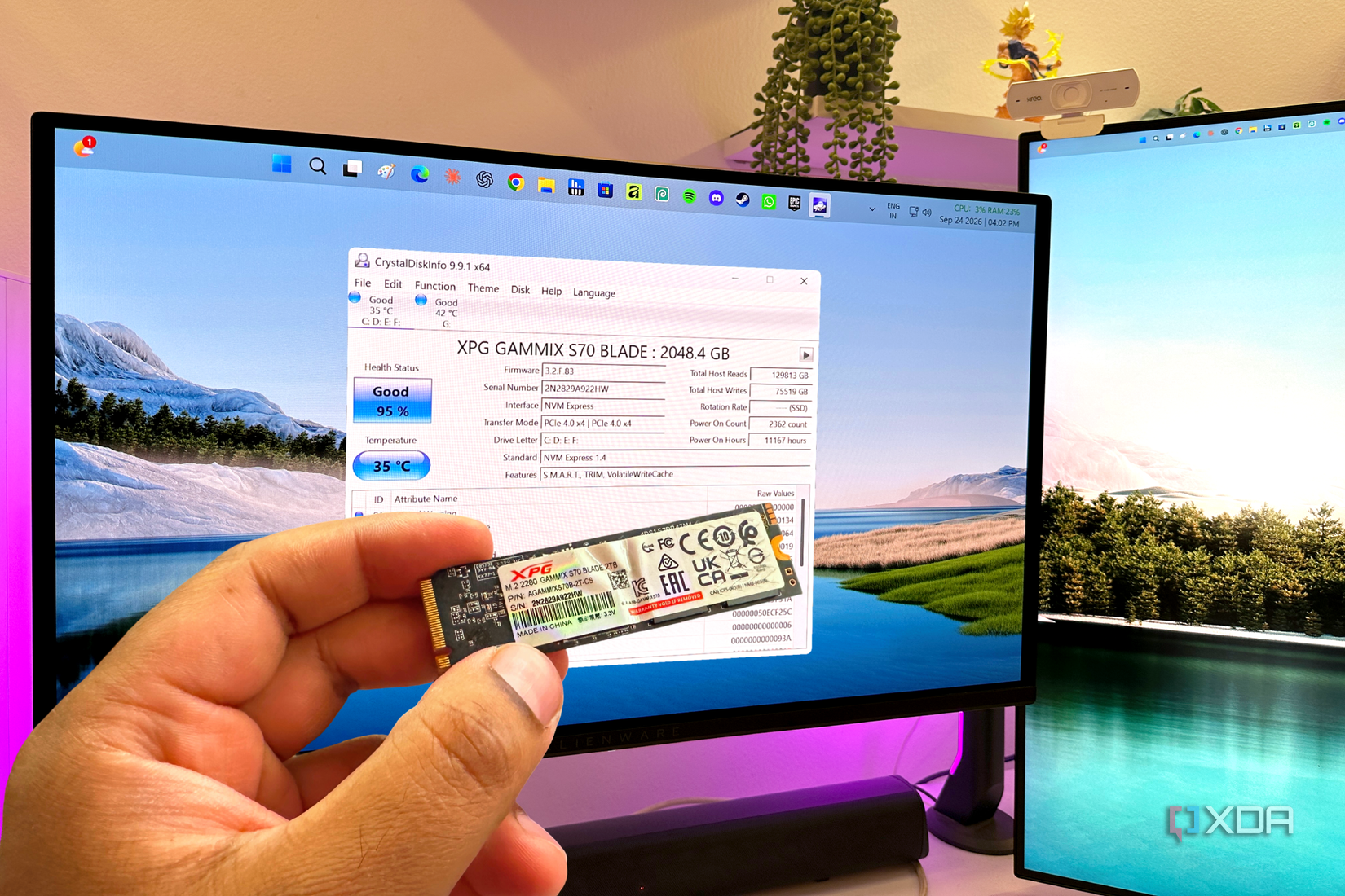 An NVMe SSD held in a hand in front of a dual-monitor setup