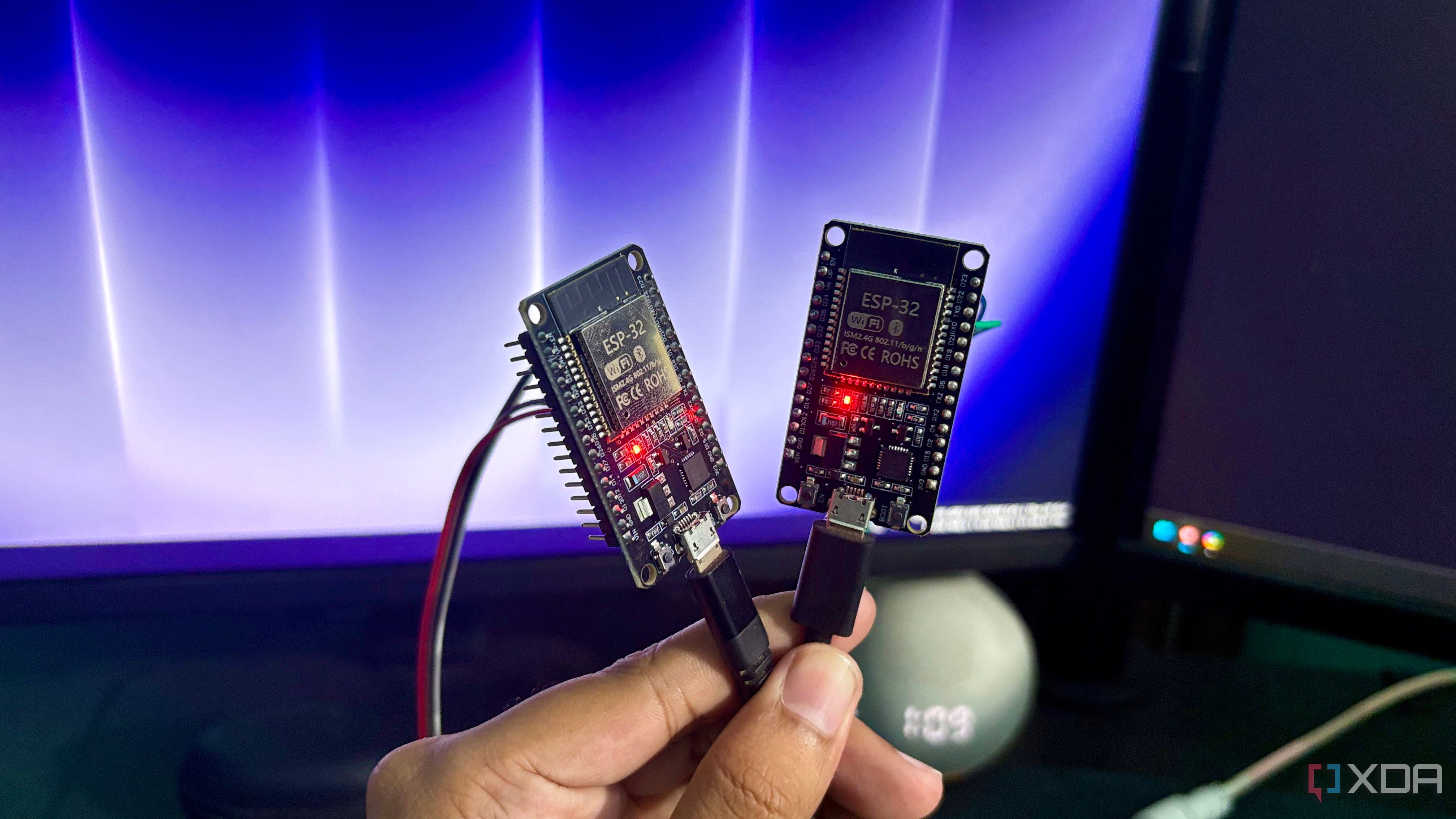 A hand holding two powered ESP32 development boards side by side in front of a monitor