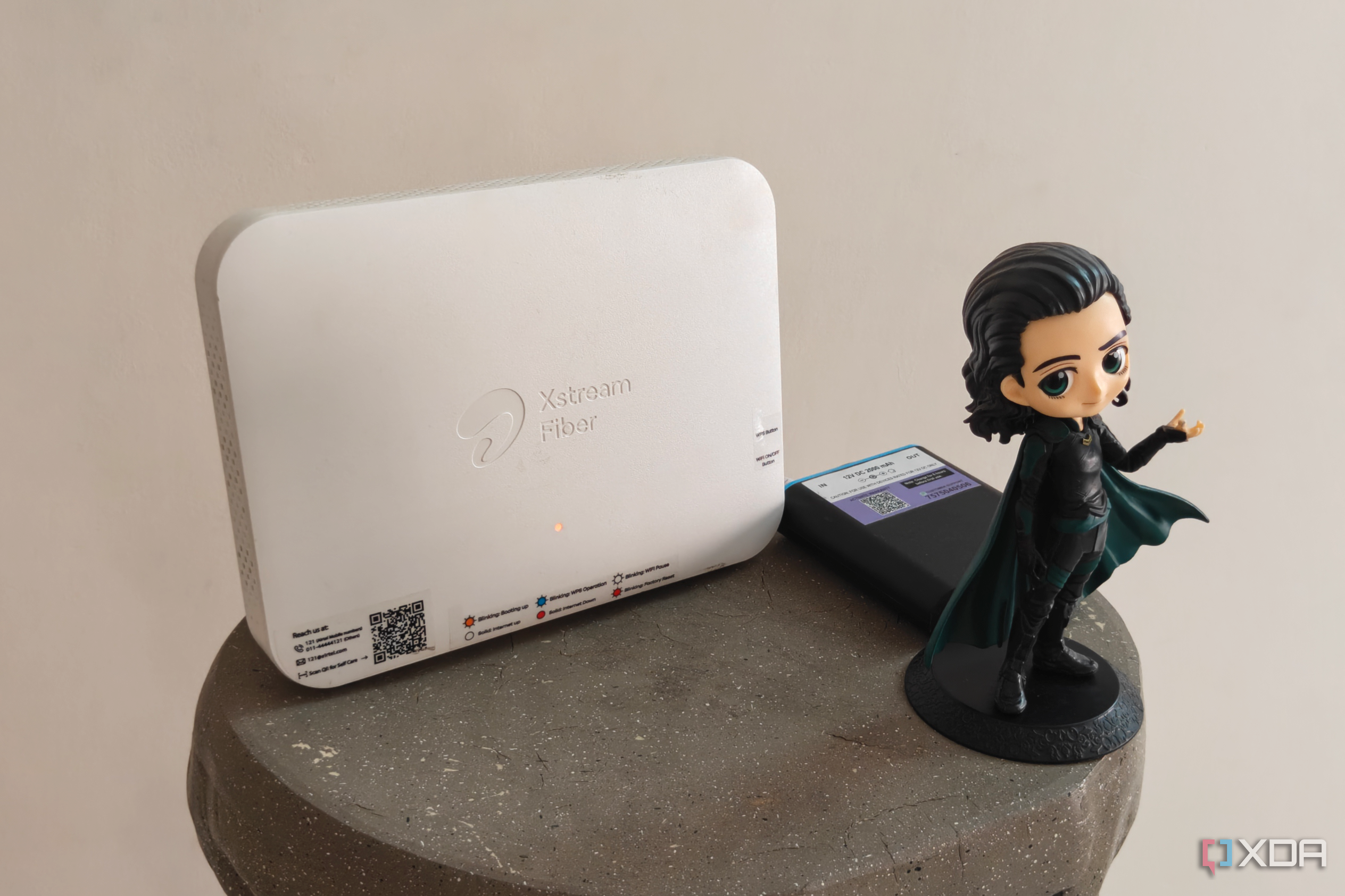 A white Wi-Fi router next to a mini UPS and a figurine on a grey surface